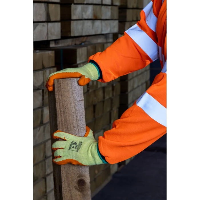 A close-up of a person wearing high-visibility orange and white work sleeves and lime green work gloves with orange fingertips, holding a wooden post in front of a stack of lumber.