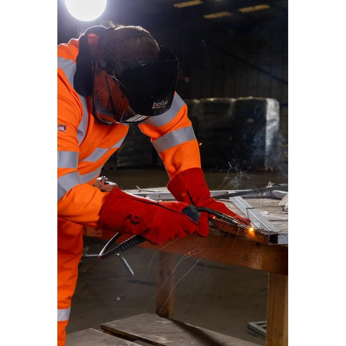 A person in a high-visibility orange welding suit with reflective stripes, a black welding helmet, and red gloves is welding metal at a workbench in a workshop. Sparks fly from the welding torch.