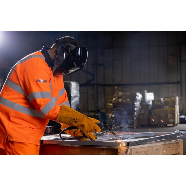 A welder in a high-visibility orange safety suit with reflective stripes, wearing a dark welding helmet and yellow gloves, actively welding metal in a dim industrial workshop setting with sparks flying.