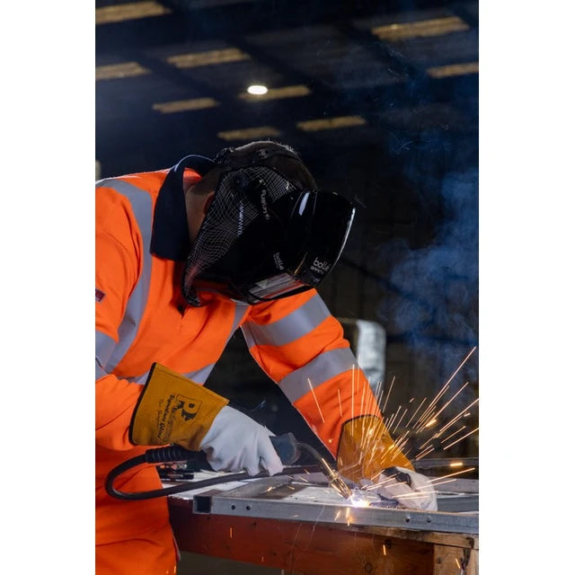 A welder in a high-visibility orange jacket and protective mask is actively welding metal, with bright sparks flying in a workshop setting.